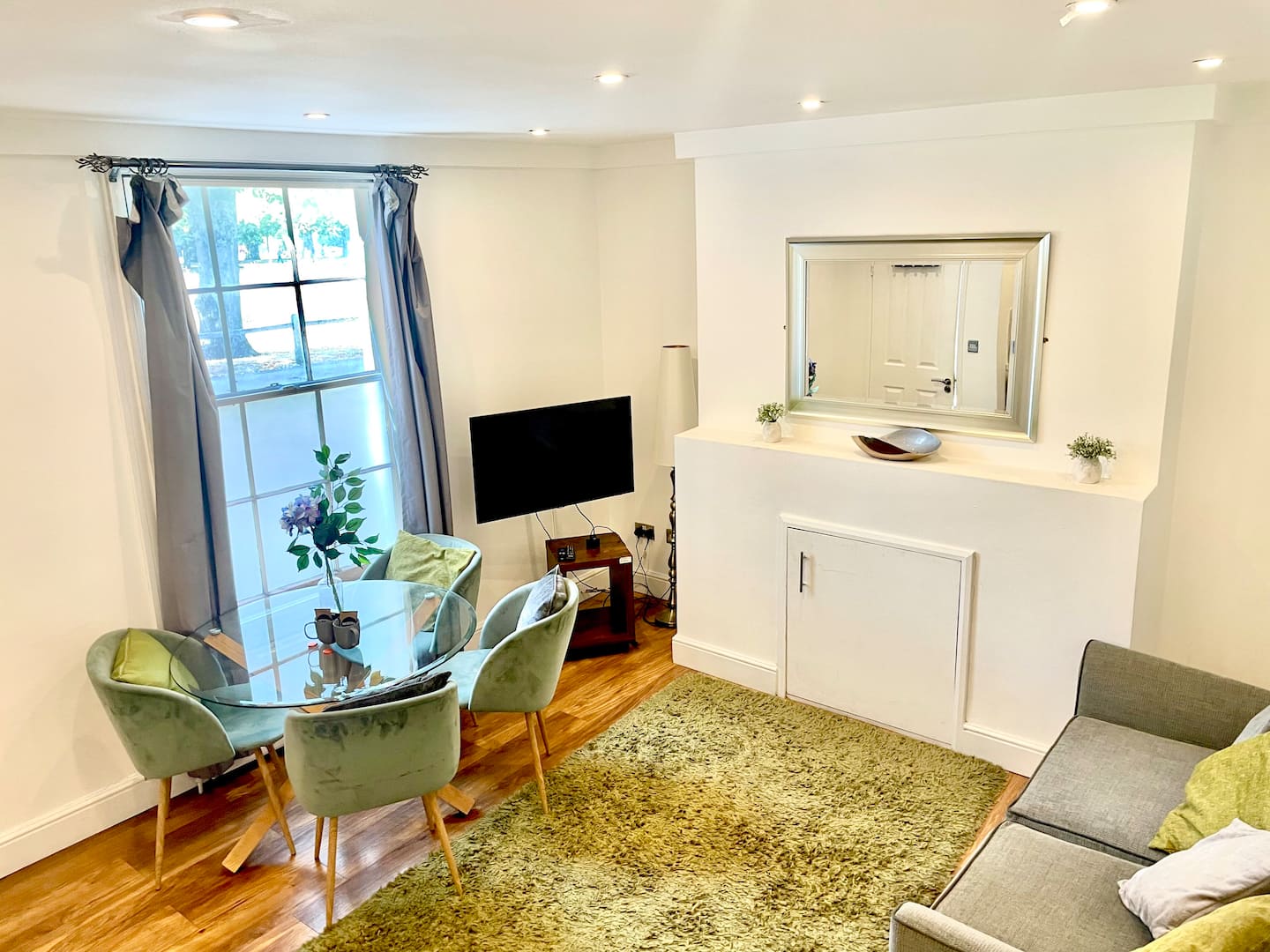 Bright living and dining area with glass table, velvet chairs and sash window overlooking the park